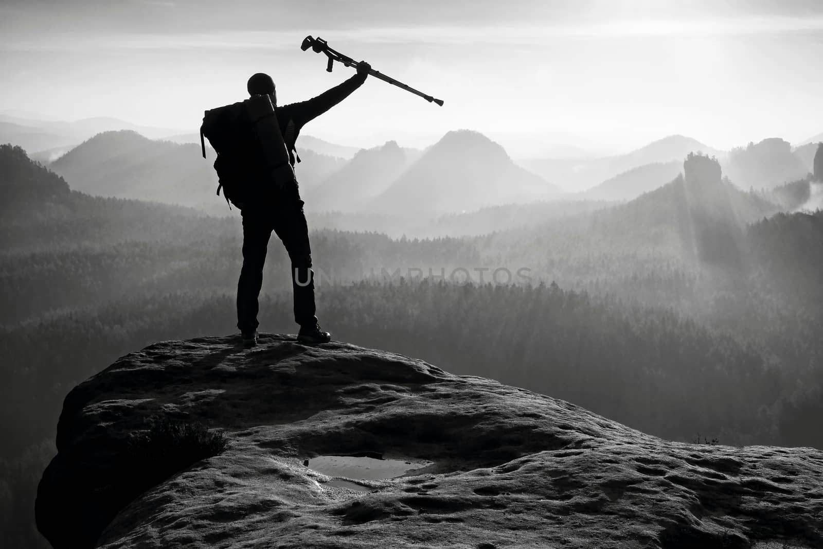 Tourist with medicine crutch above head achieved mountain peak. Hiker with broken leg in immobilizer. Deep misty valley bellow silhouette of man with hand in air. Spring daybreak