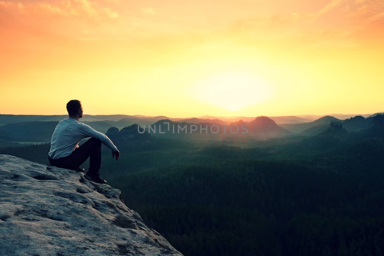 Young hiker in black trousers and grey shirt  sit on cliff's edge. Tourist watching sunrise above  misty hilly valley bellow mountain