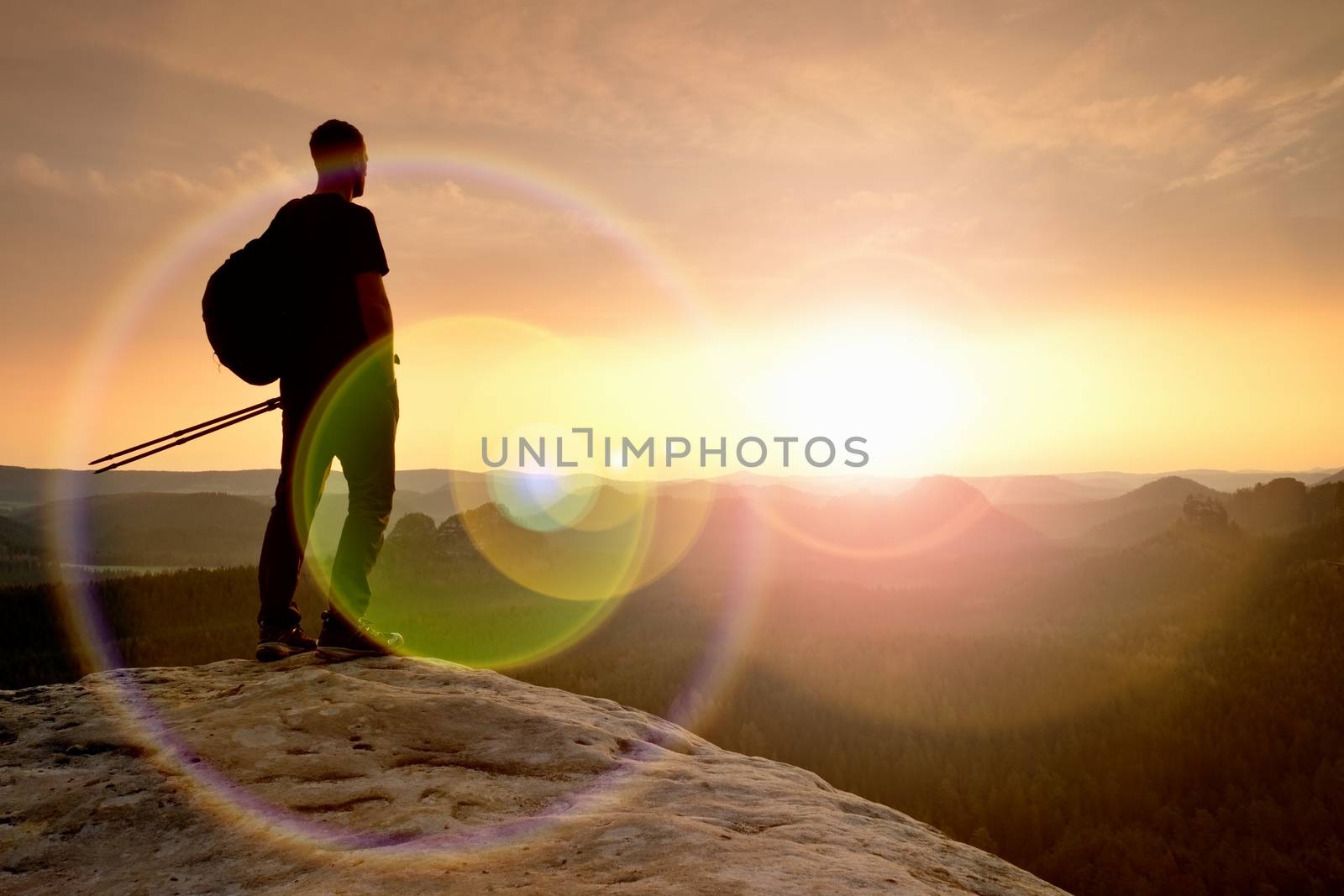 Thinking hiker in black on rocky peak. Wonderful daybreak. Ginger hair man in black at the end of sharp cliff above deep valley. Strong lens defect, reflection