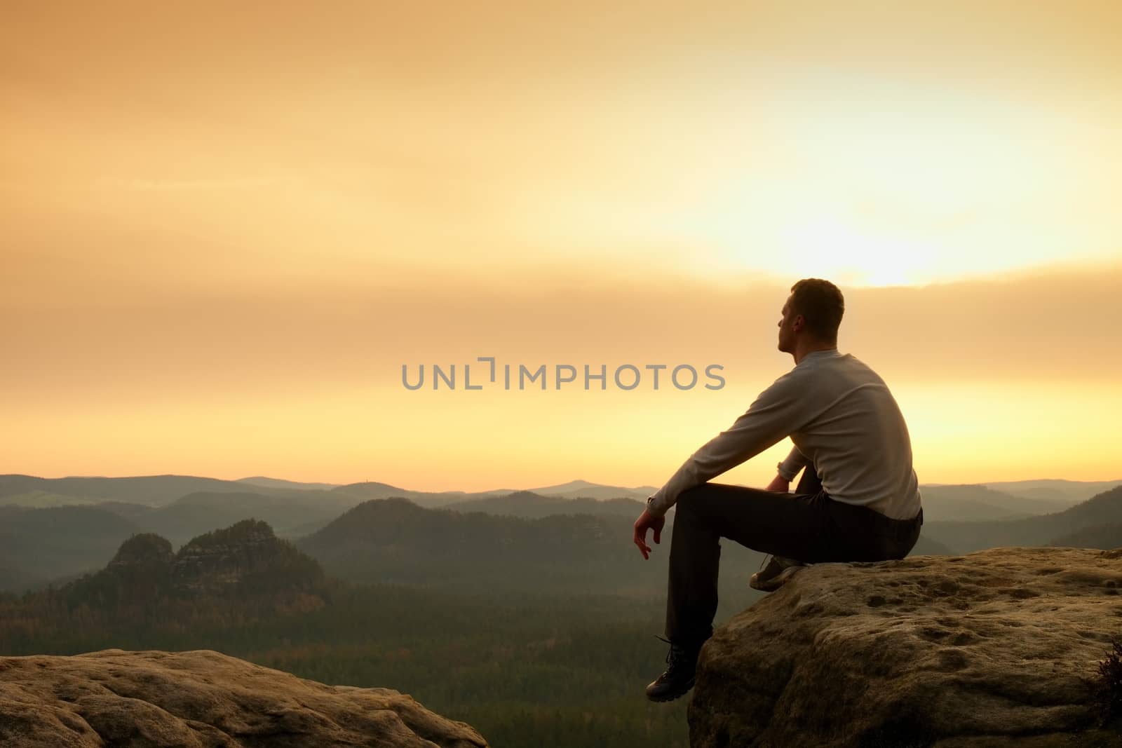 Hiker in black sit alone on the rock summit. Wonderful daybreak in mountains, heavy mist in deep valley. Man sit on the rock.