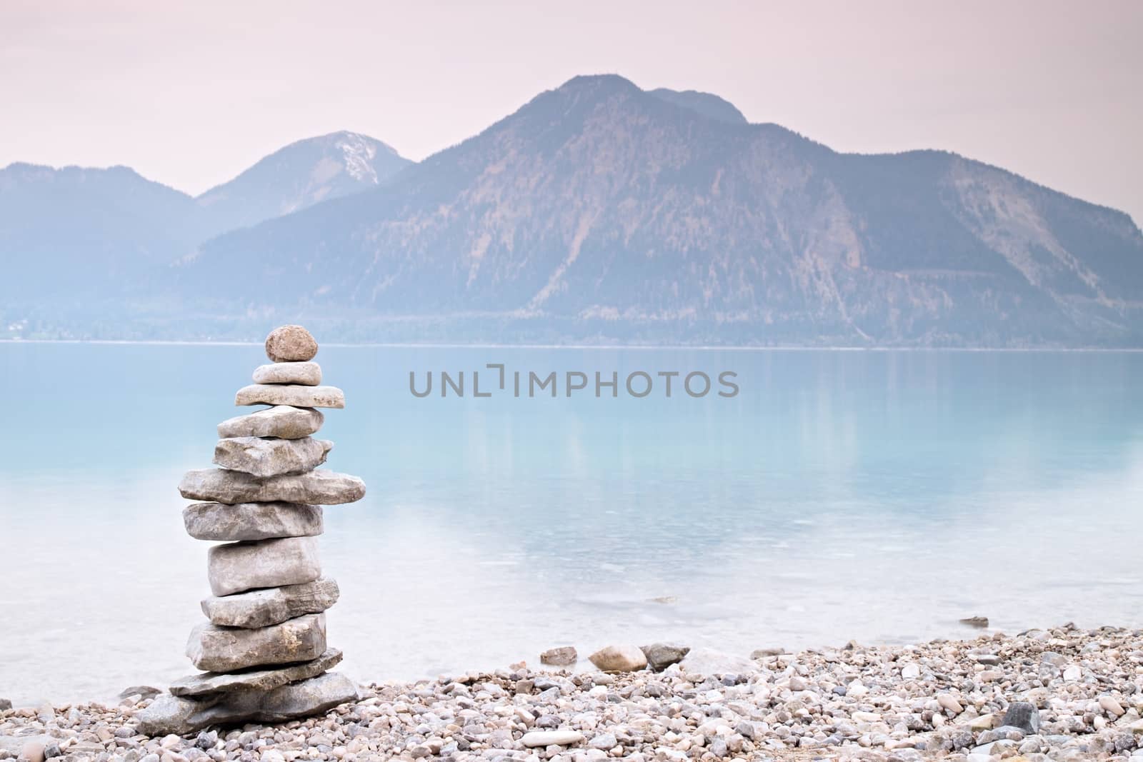 Balanced stone pyramide on shore of blue water of mountain lake. Blue mountains in water level mirror. Children built pyramid from pebbles.  Poor lighting conditions.