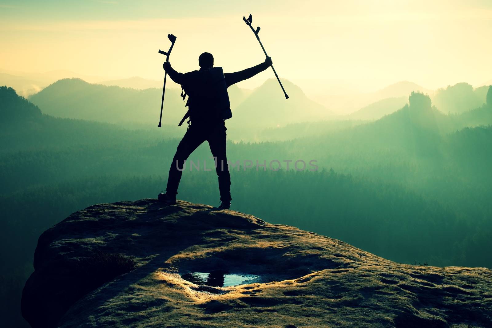 Hiker with broken leg in immobilizer. Tourist with  medicine crutch above head achieved mountain peak. Deep misty valley bellow silhouette of man with hand in air. Spring daybreak