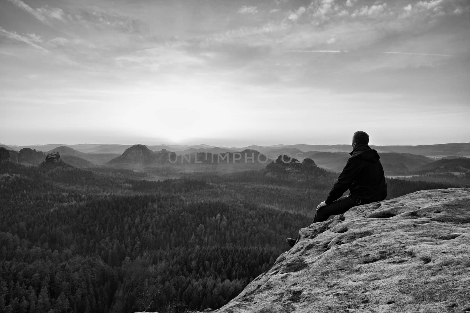 Young hiker in black and shirt is sitting on cliff's edge and enjoing spring morning in nature. View into misty hilly valley bellow