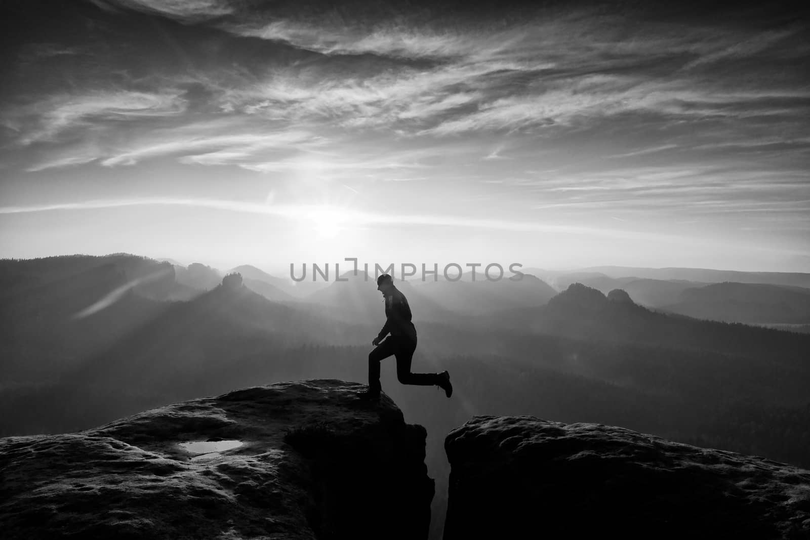 Crazy hiker in black is jumping between rocky peaks. Happy man. Wonderful daybreak in rocky mountains, heavy orange mist in deep valley. Miracle of nature