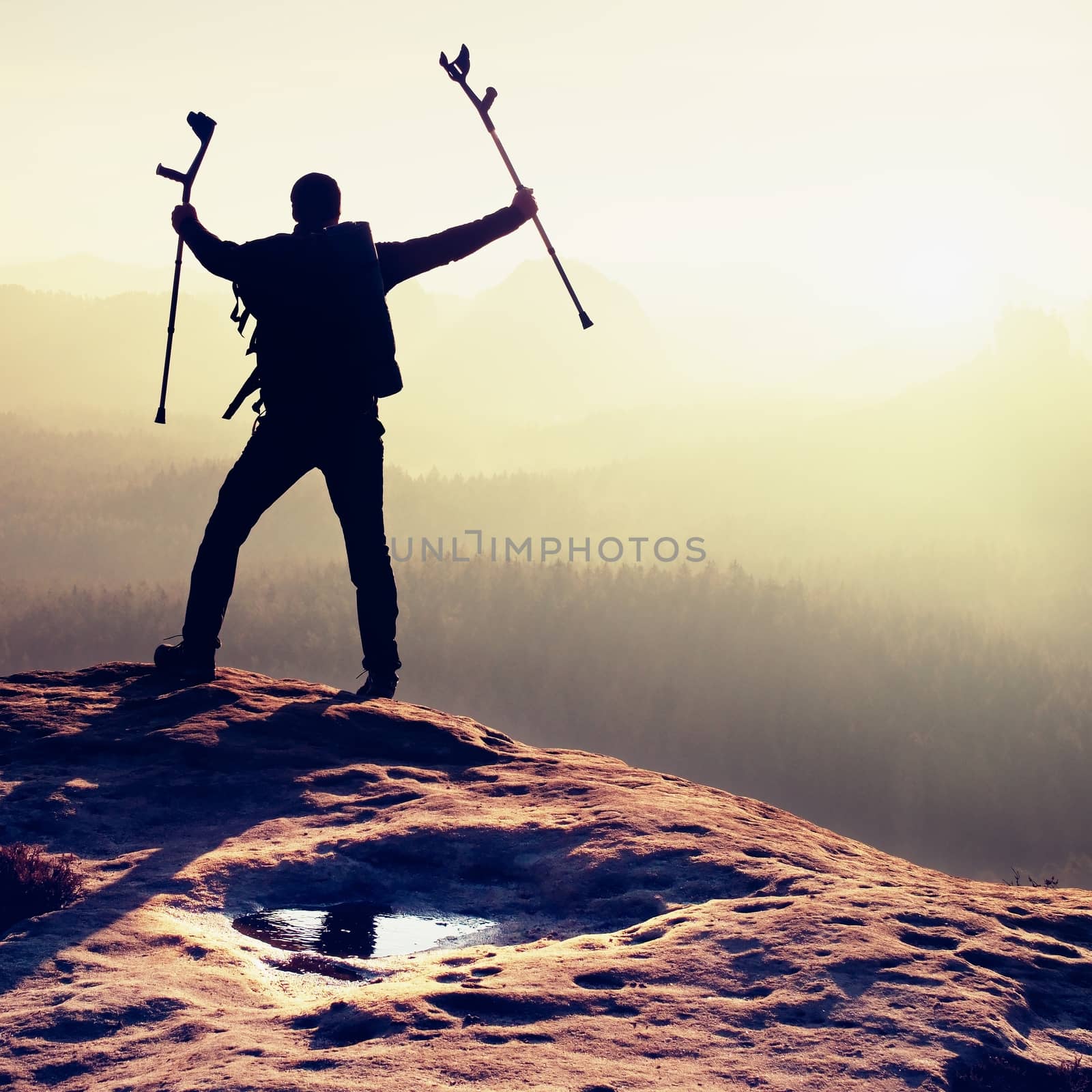 Tourist with  medicine crutch above head achieved mountain peak. Hiker with broken leg in immobilizer.  Deep misty valley bellow silhouette of man with hand in air. Spring daybreak