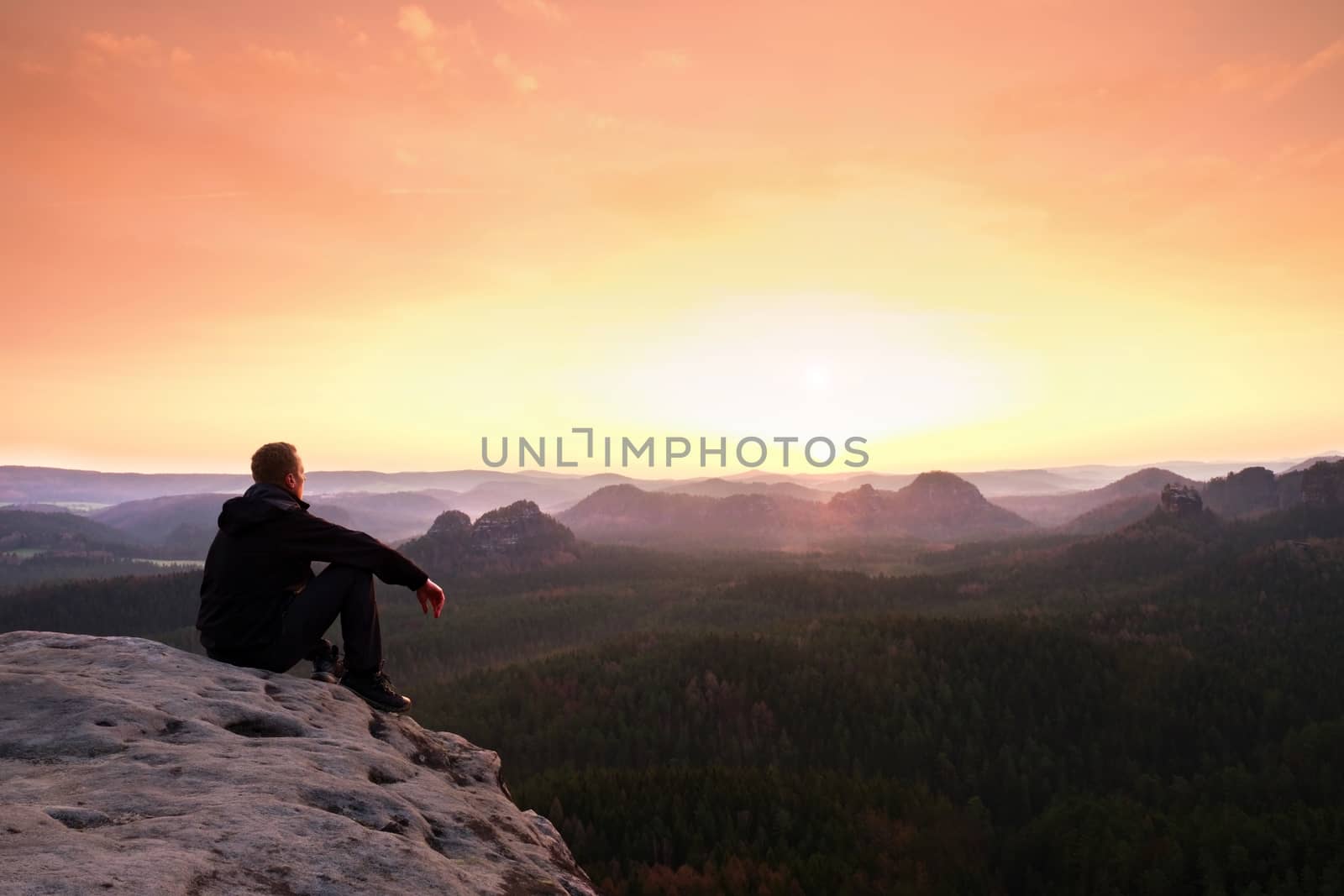 Adult tourist in black  sit on cliff's edge. Man enjoying morning  and looking to orange daybreak at misty horizon