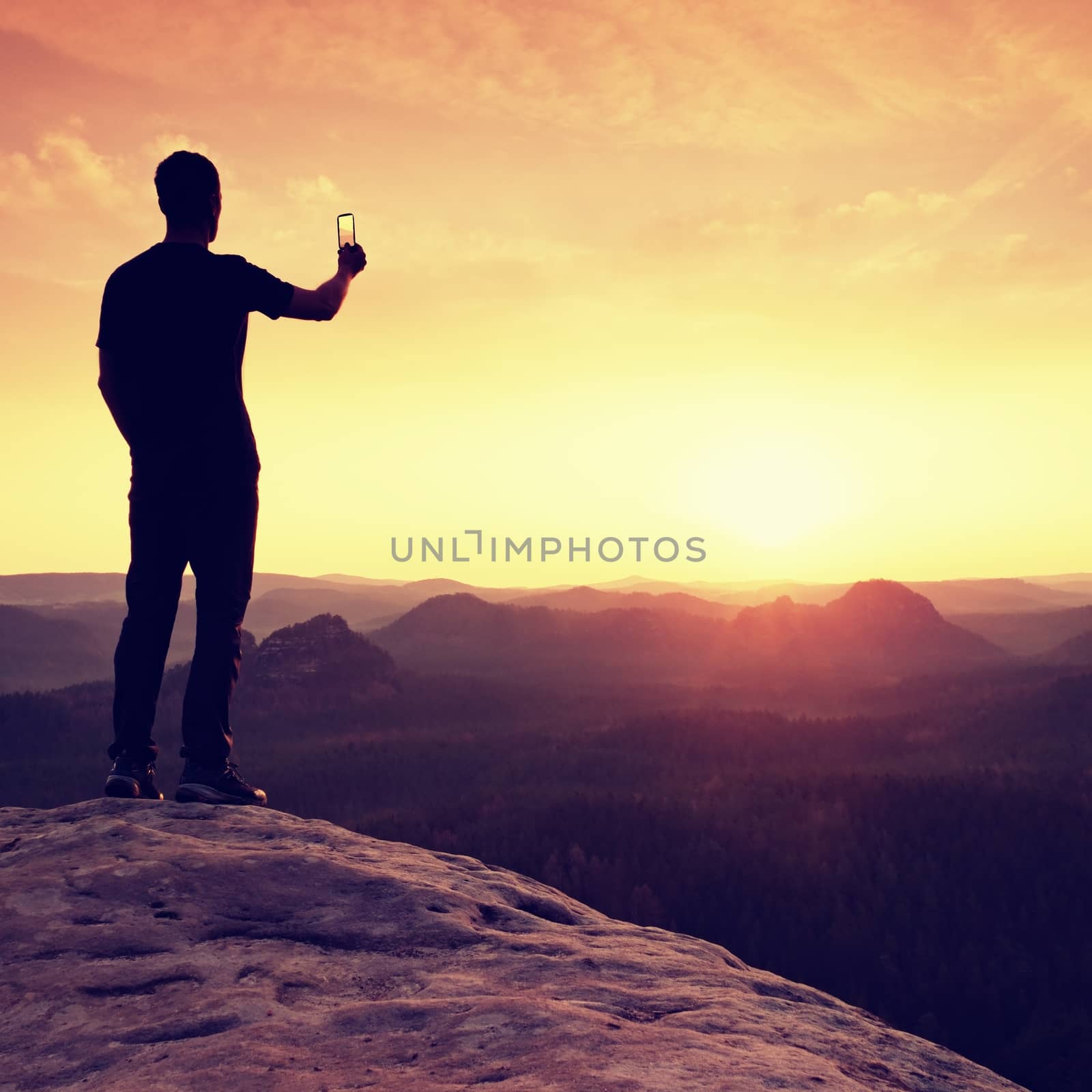 Man takes photos with phone on rock empire. Dreamy fogy mountains, spring orange pink misty sunrise in a beautiful valley of rocky mountains.