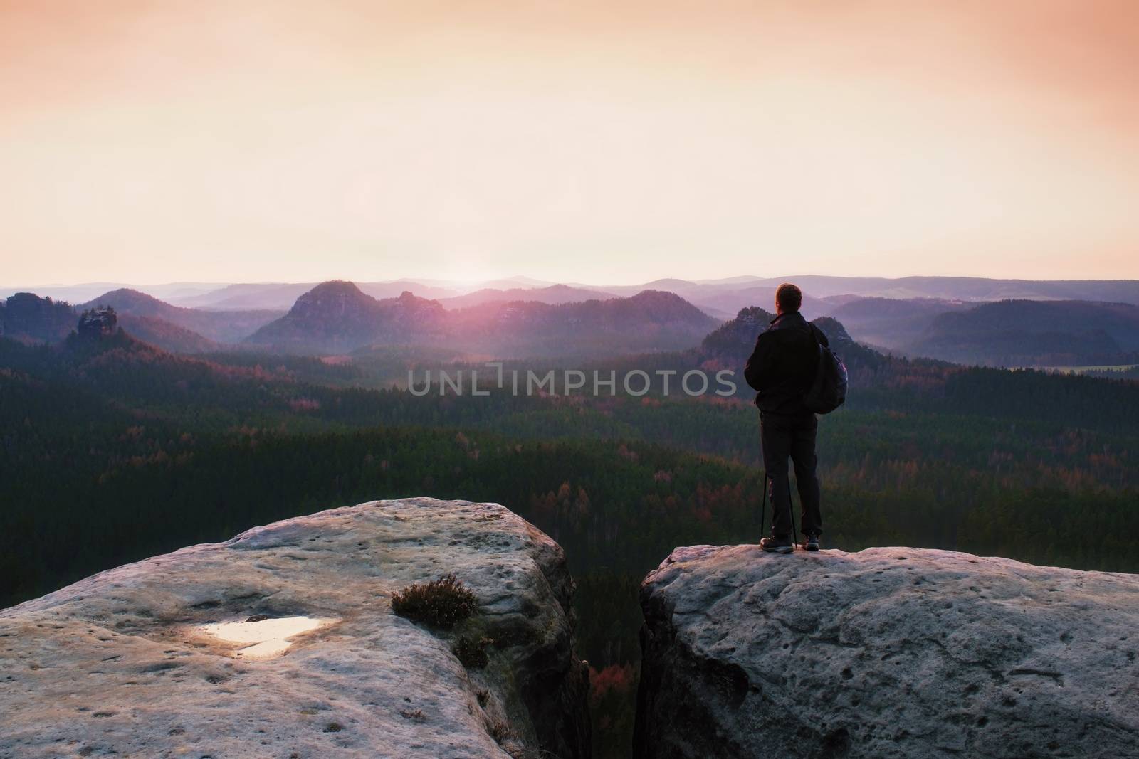 Tall tourist guide on rock with pole in hand. Hiker with sporty backpack stand on cliff above misty valley. Sunny spring daybreak in rocky mountains.