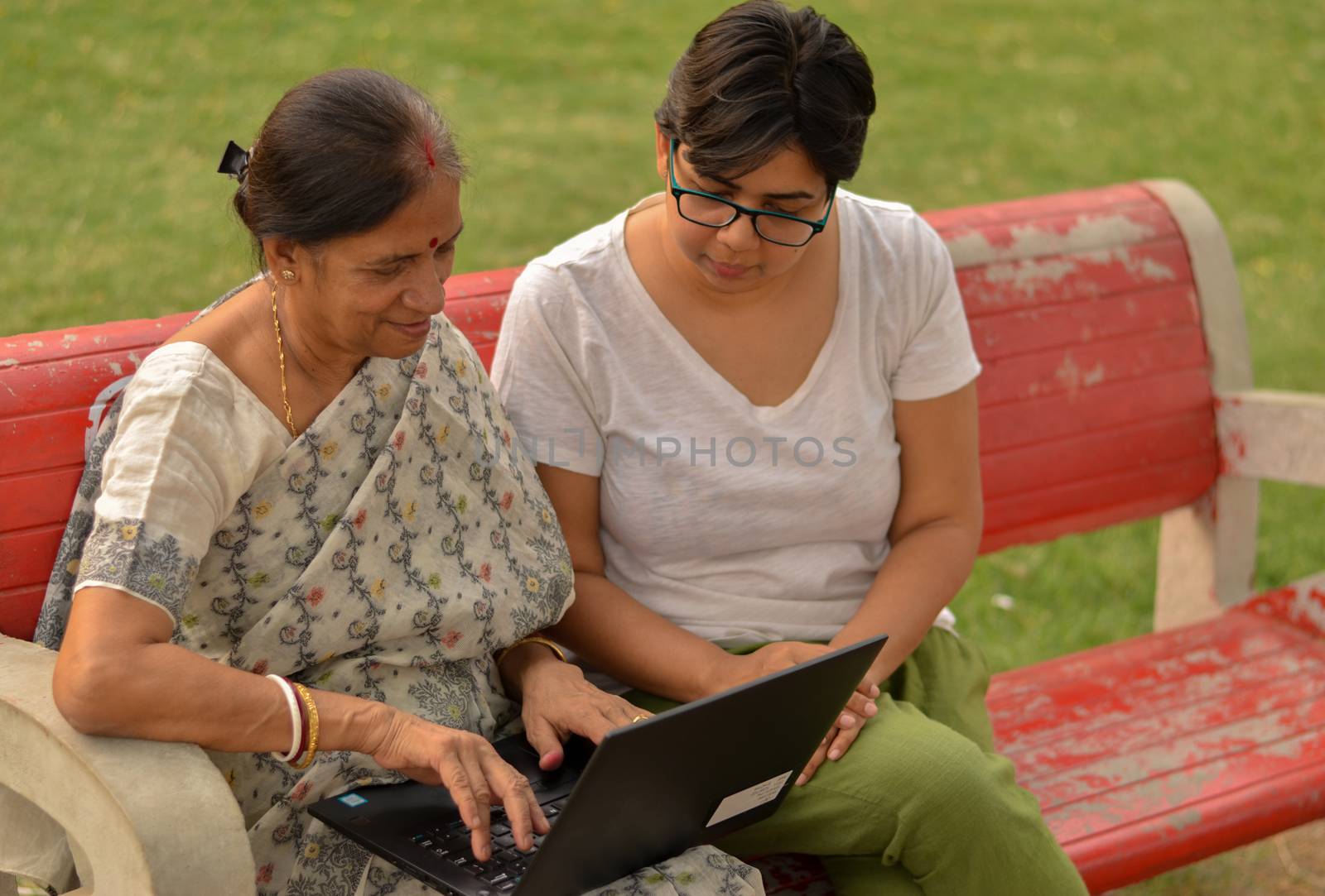 Side view of young Indian girl helping an old Indian woman on a laptop sitting on a red bench in a park in New Delhi, India. Concept Digital literacy / Education