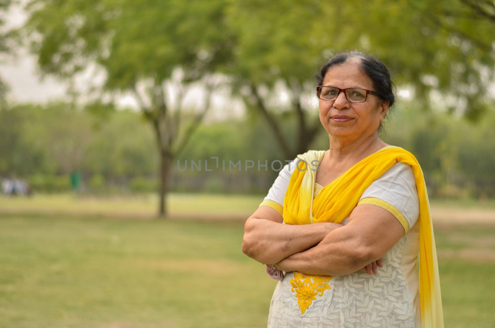 Smart senior north Indian woman standing, posing for the camera with hands crossed in a park wearing yellow salwar kameez in summers in New Delhi, India