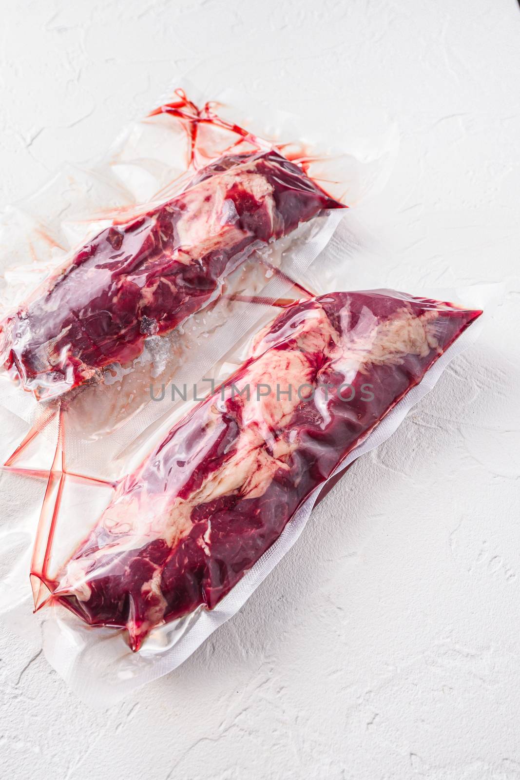 Pair of chuck roll beef steak, vacuum packed organic meat for sous vide cooking on white concrete textured background, side view. by Ilianesolenyi