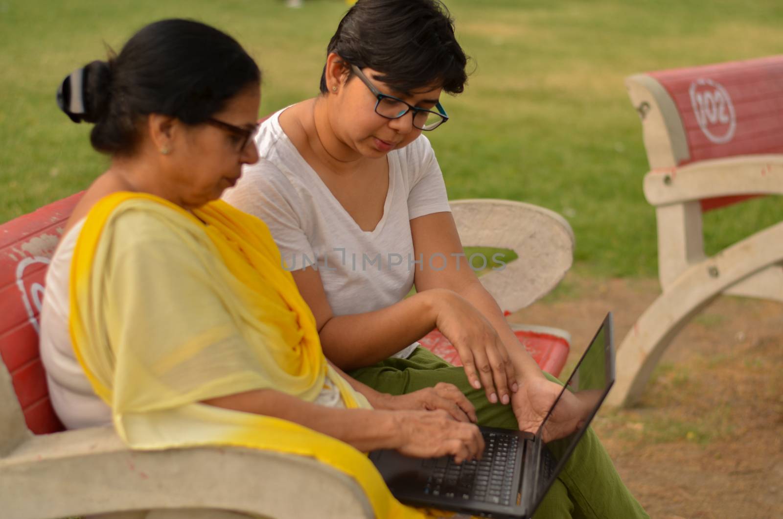 Side view of young Indian girl helping an old Indian woman on a laptop sitting on a red bench in a park in New Delhi, India. Concept Digital literacy / Education by jayantbahel