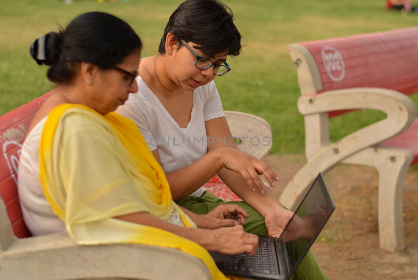 Side view of young Indian girl helping an old Indian woman on a laptop sitting on a red bench in a park in New Delhi, India. Concept Digital literacy / Education by jayantbahel