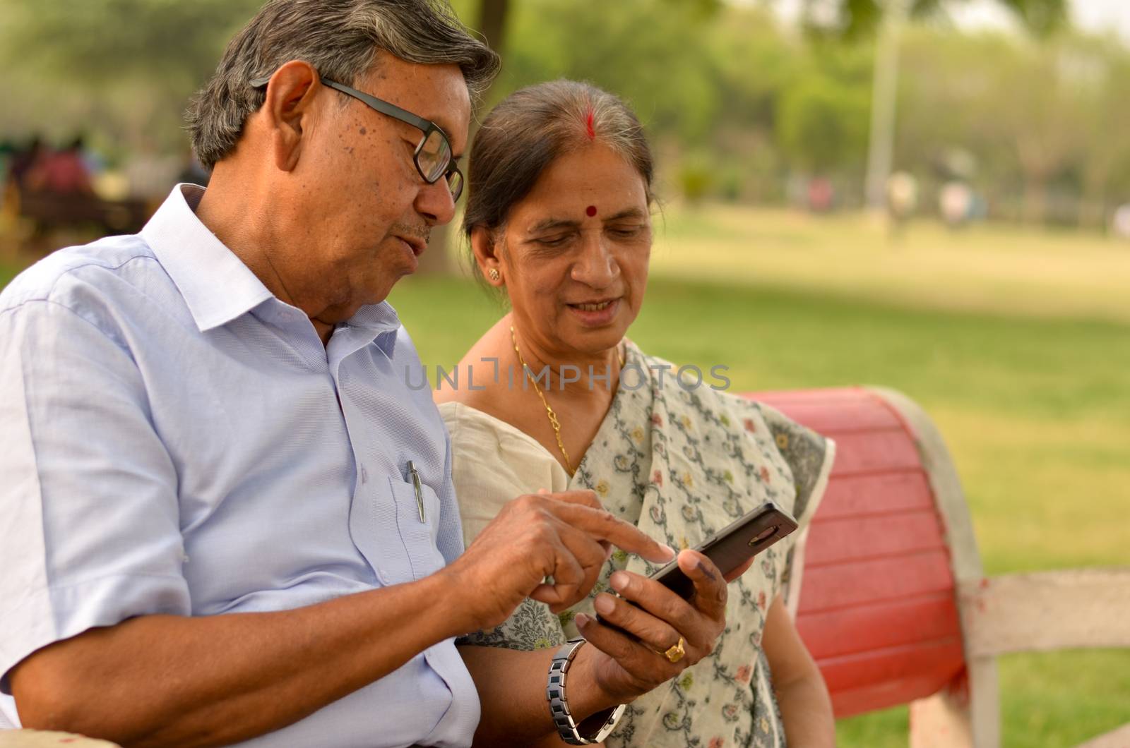 Portrait of senior couple sitting in park bench and looking at their smart phone and laughing in New Delhi, India with focus on the hands