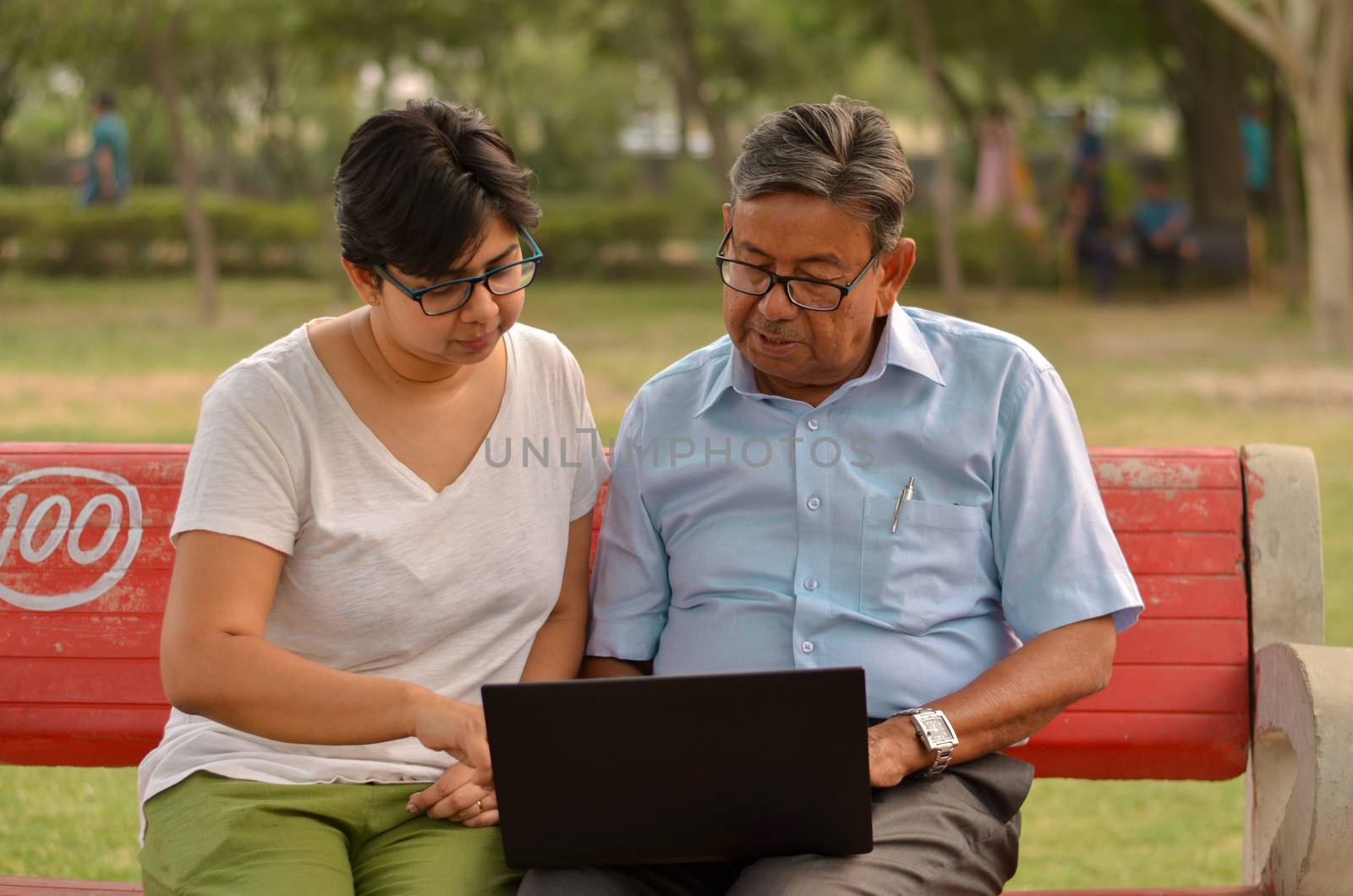 Young Indian woman manager in western formals / shirt helping old Indian man on a laptop promoting digital literacy for elderly in a park in New Delhi, India.Concept: father daughter/ Digital literacy