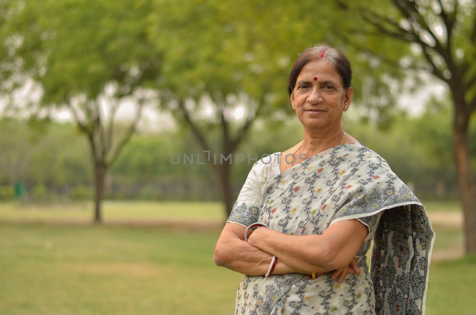 Smart senior north Indian woman standing, posing for the camera with hands crossed in a park wearing white/grey saree in summers in New Delhi, India