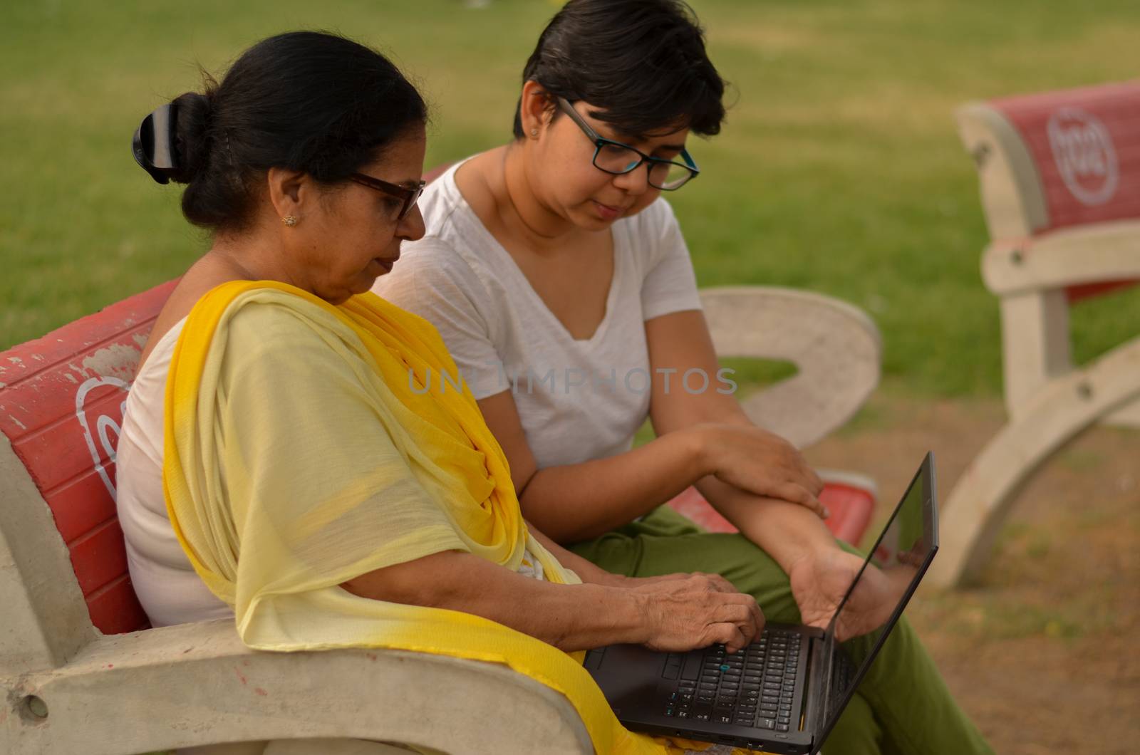 Side view of young Indian girl helping an old Indian woman on a laptop sitting on a red bench in a park in New Delhi, India. Concept Digital literacy / Education