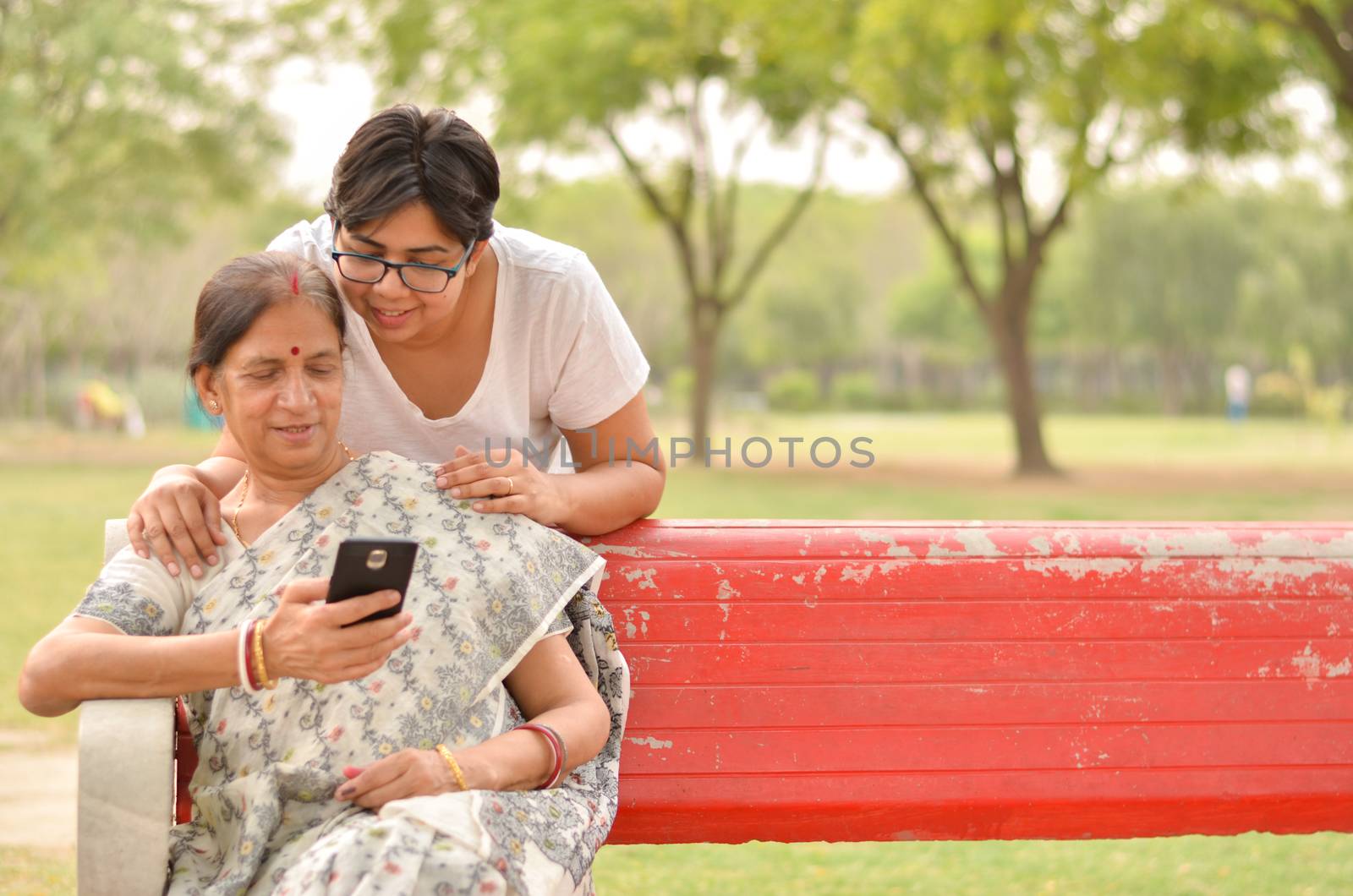 Young Indian girl keeping her hands on her old Indian Bengali mother's shoulders looking at the mobile phone and busy talking sitting on a red bench in a park in New Delhi, India. Concept Mother's day