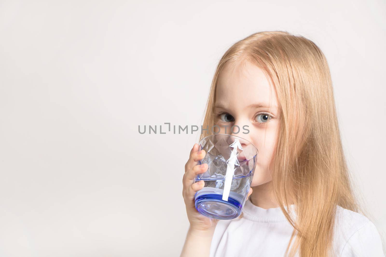 A beautiful girl of Caucasian appearance drinks water from a glass on a white background. A young blonde model girl holds a glass of water.