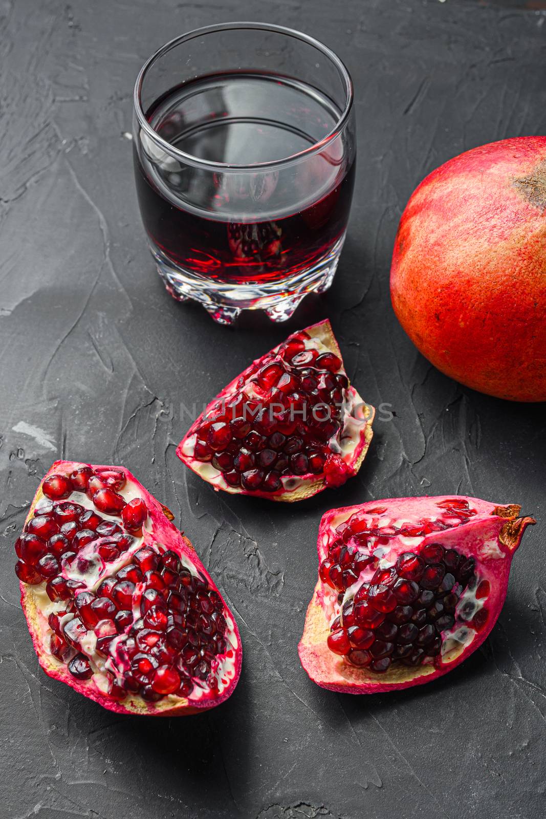 Pomegranate cuts with seeds in front of garnet cut and juice in glass side view, on black background, selective focus by Ilianesolenyi