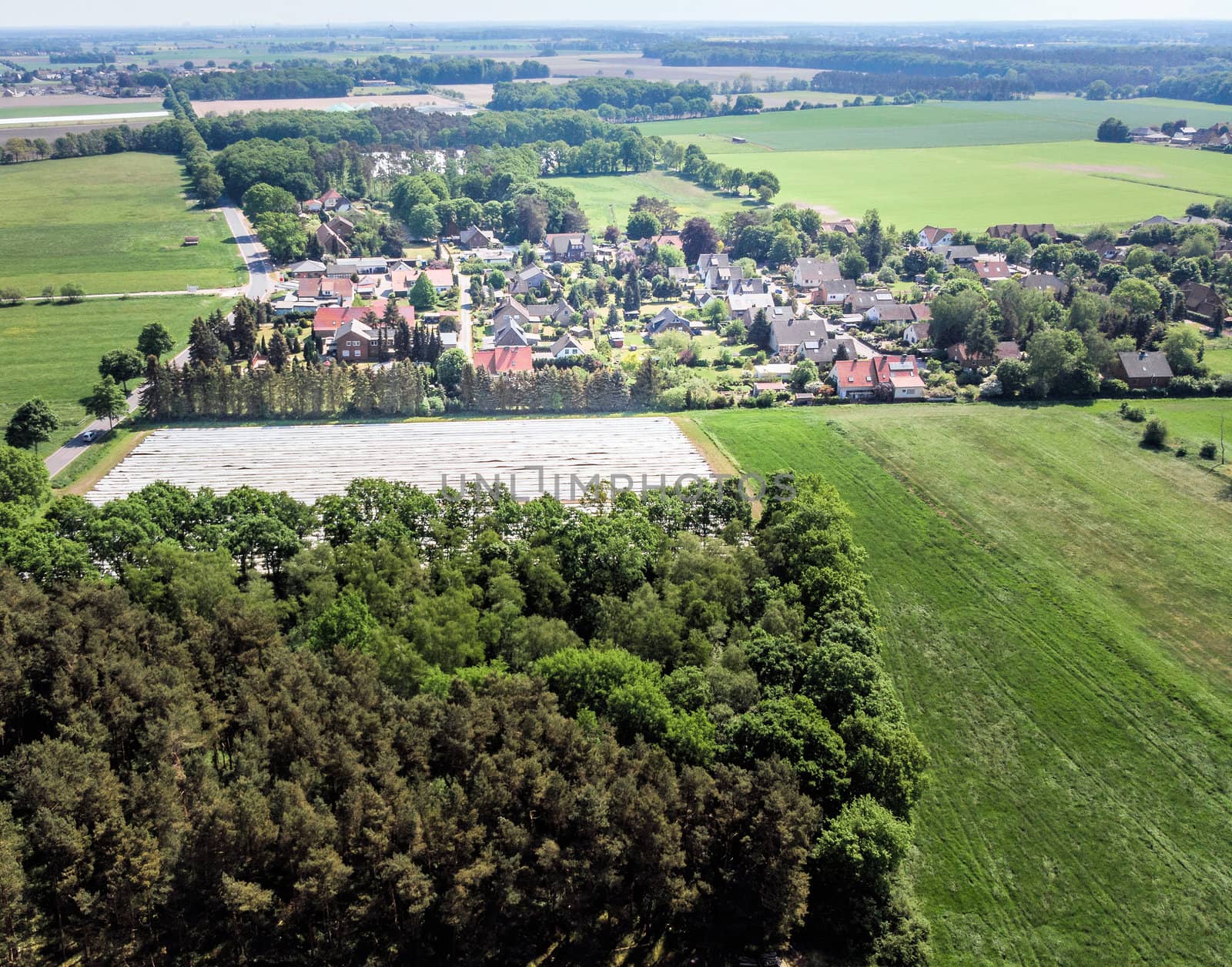 Aerial view of a small village in the distance behind a piece of forest and an asparagus field covered with foil by geogif