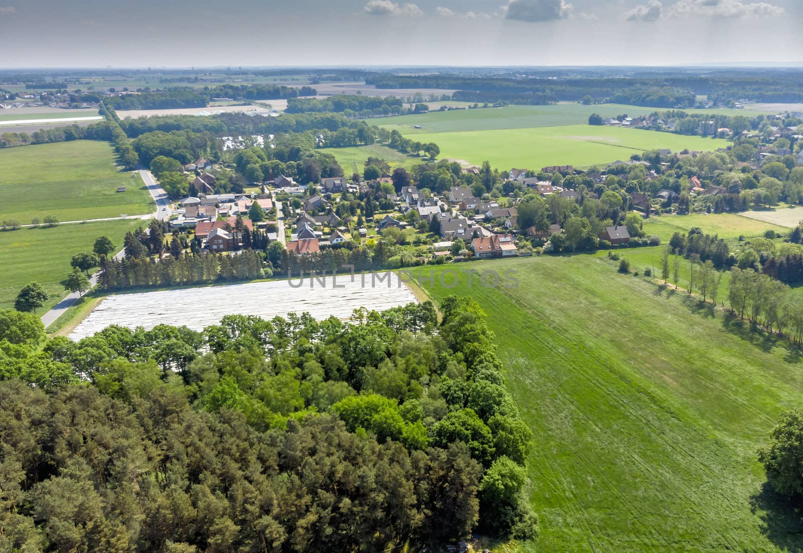 Aerial view of a small village in the distance behind a piece of forest and an asparagus field covered with foil by geogif