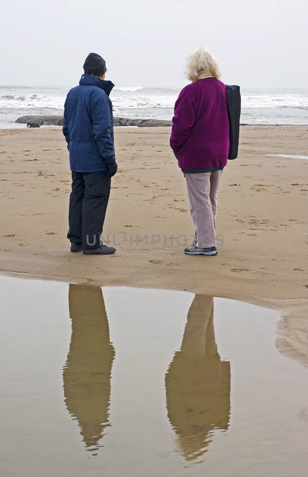Two people stood on the beach in winter with a reflection in a small pool