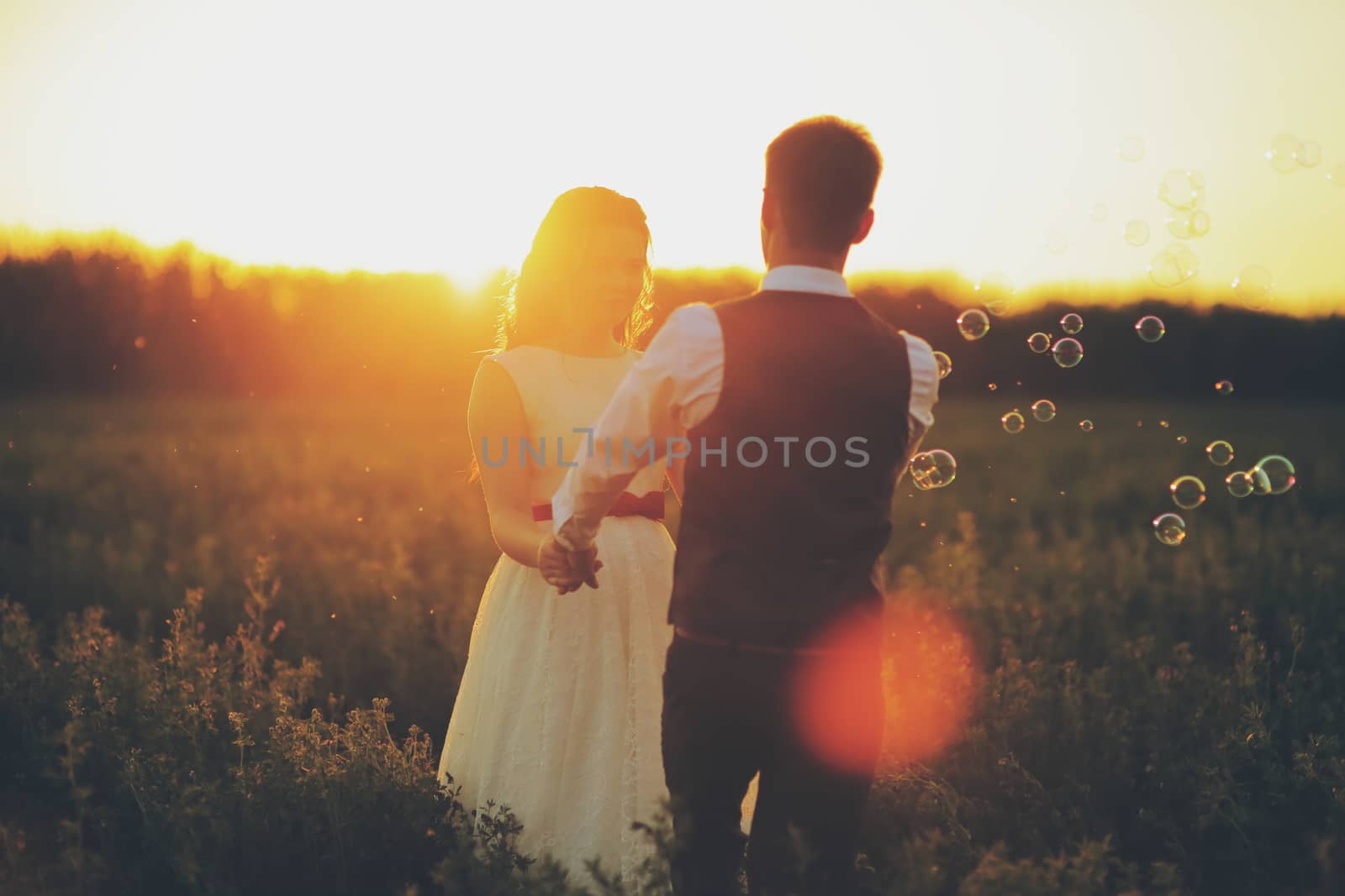 The bride and groom hold hands dancing in the park. Sunset light. Soap bubbles. Wedding. Happy family concept. by selinsmo