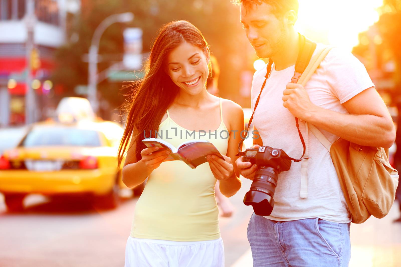 Travel tourist couple traveling in New York reading guide book standing with SLR camera at sunset on Manhattan with yellow taxi cab in the background. Happy young multiracial couple on summer holidays