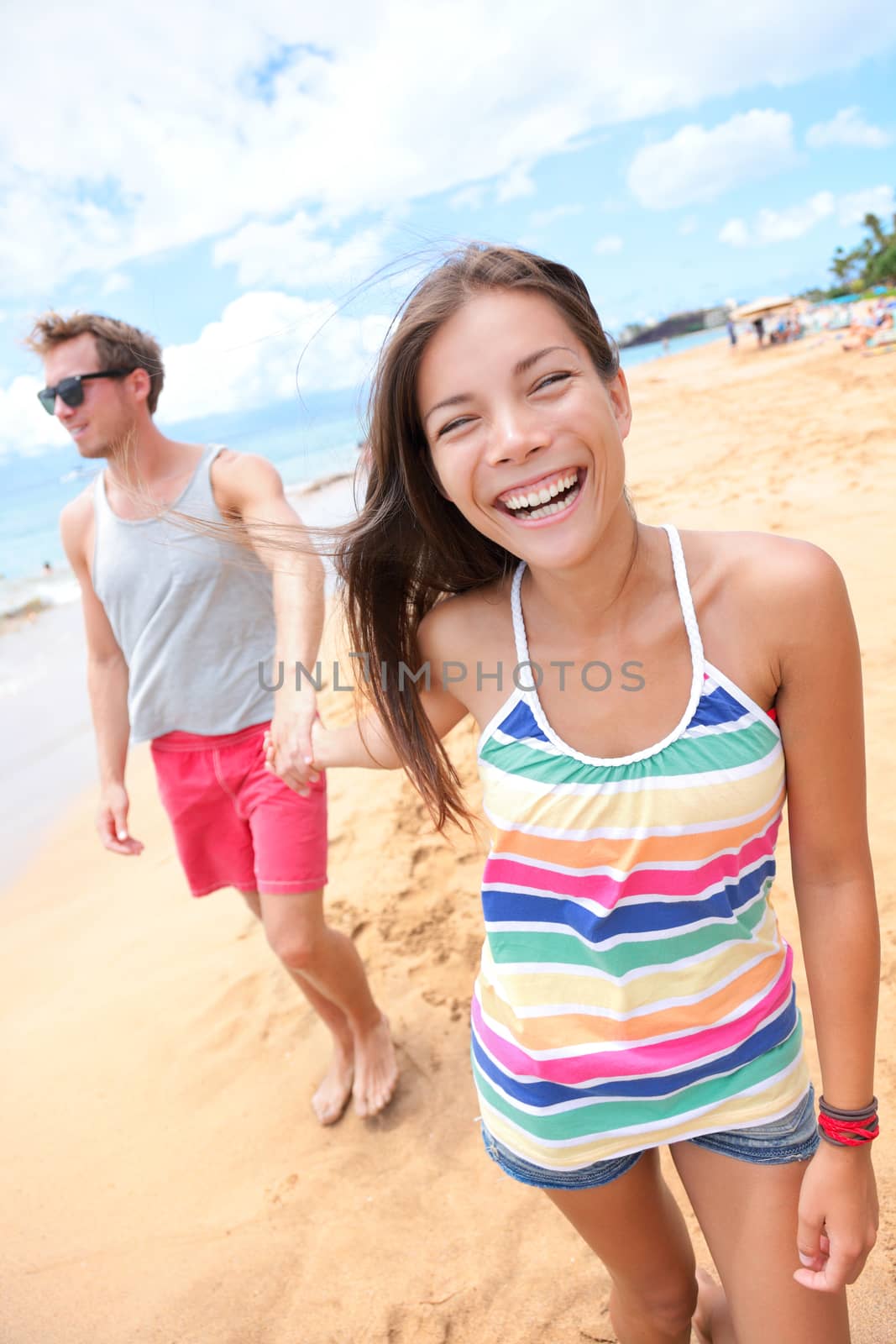 Beach people. Young interracial happy playful couple holding hands walking on beach having fun on travel vacation - woman smiling laughing man in background . Asian woman, Caucasian man on Big Island, Hawaii, USA.
