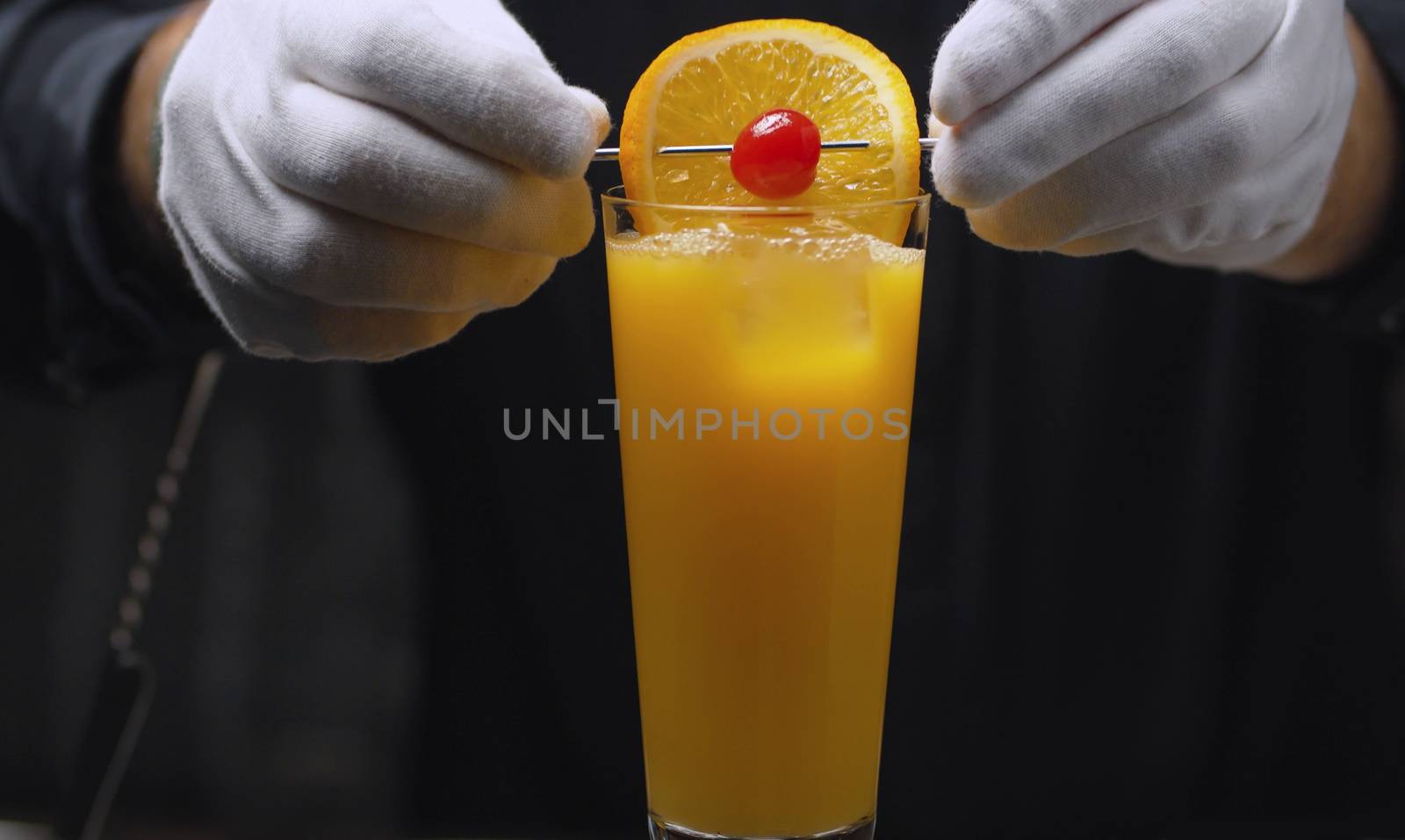 Preparing a Tequila Sunrise Cocktail. Close up bartenders hands garnish it with orange and cherry on skewer.