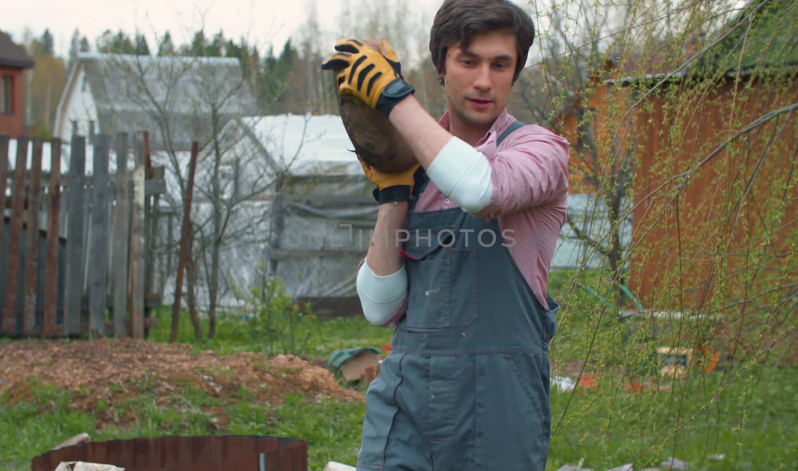 Young man carrying big stone in his hands in the courtyard. The arrangement of the yard of a country house, concept nature friendly people and a healthy active lifestyle