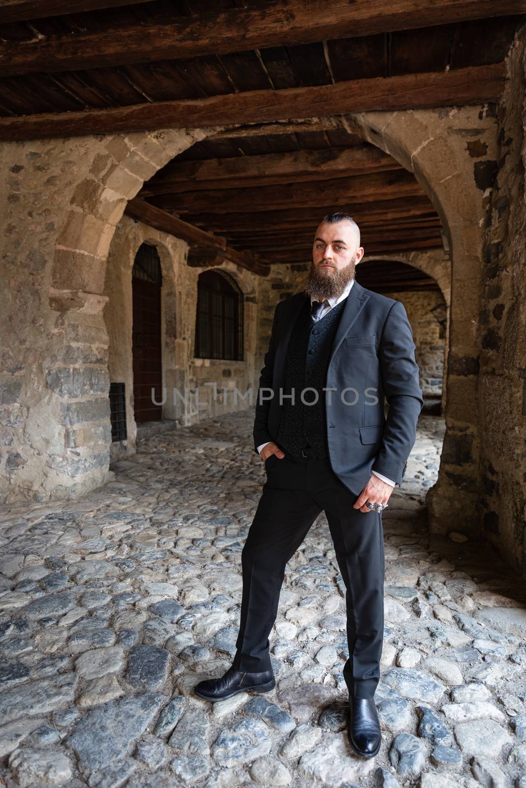 Man in dark elegant suit posing under the arcades of a medieval Italian village, full-length male portrait