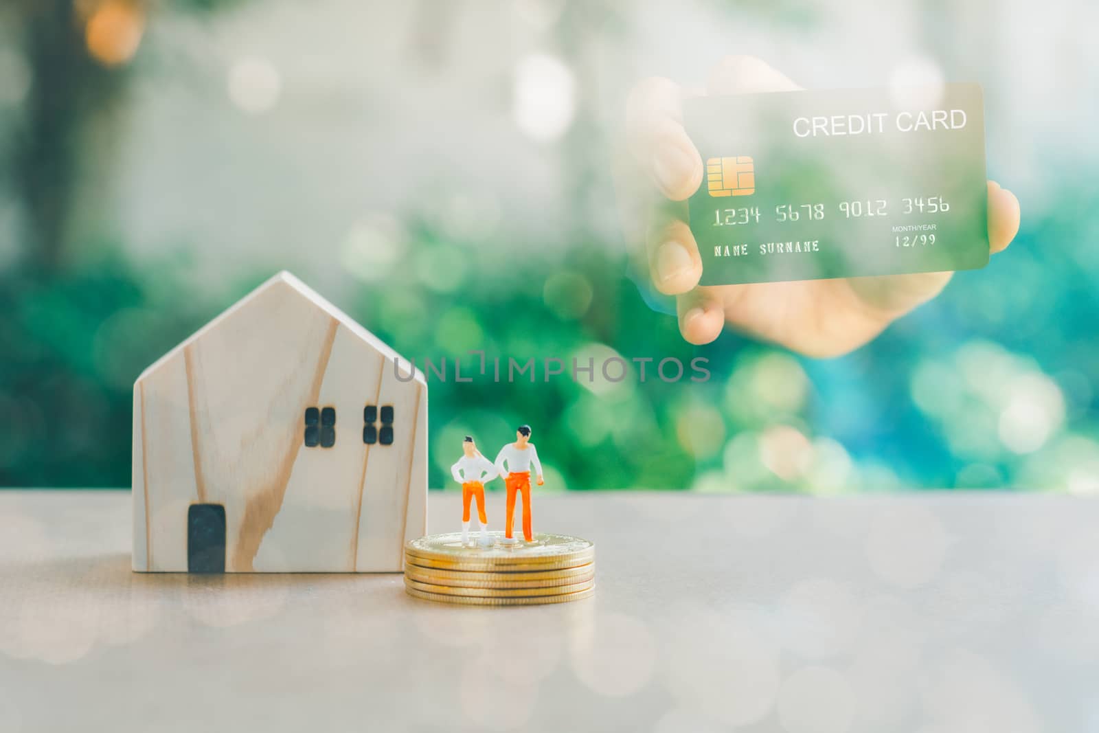 Business and financial concept. Double exposure hand holding credit card and wooden home with small people model stand on a stack of golden coins.