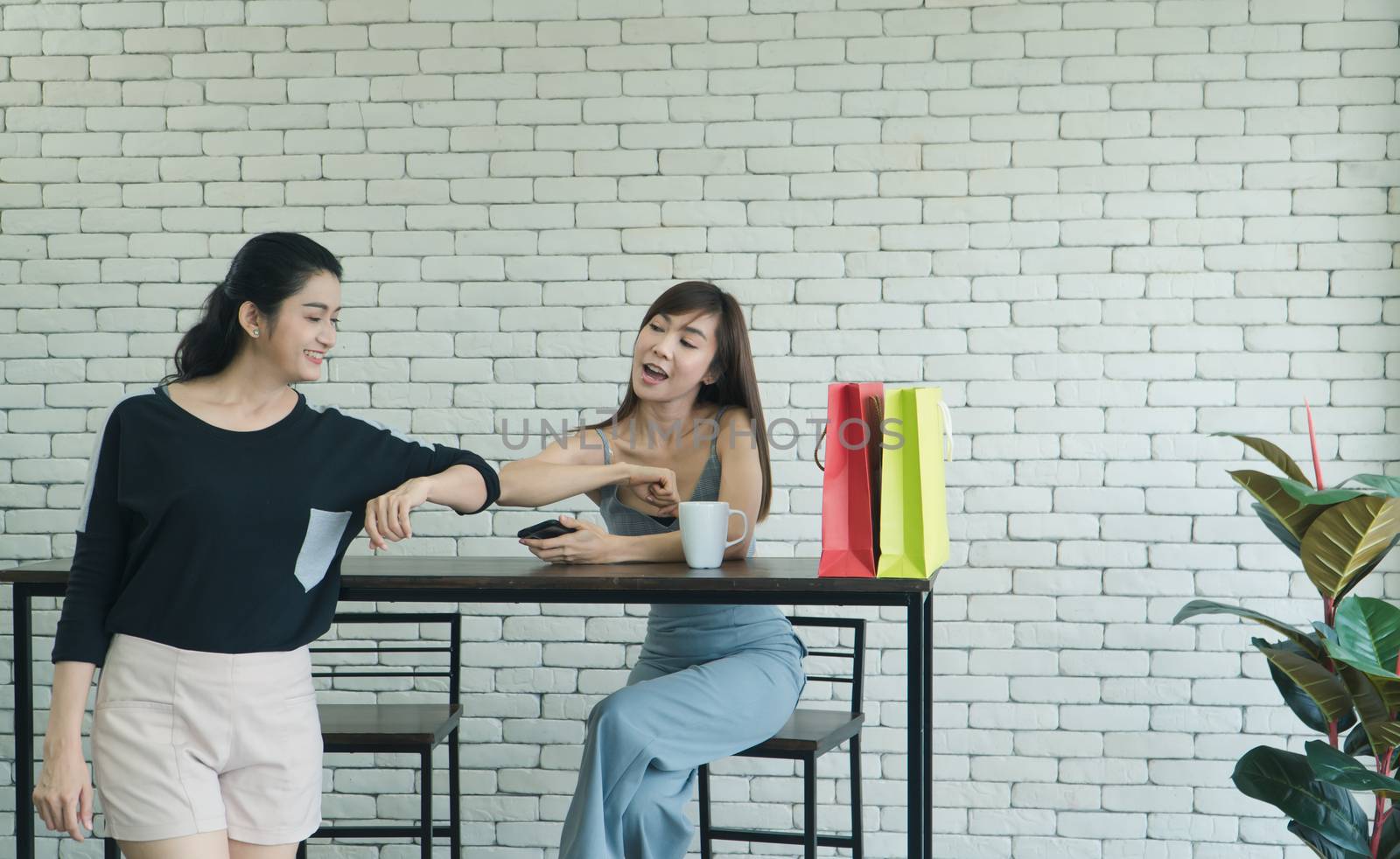 Two Asian women who are laughing and attractive are friends, talking about coffee in a cafe. Beautiful and confident women use their smartphones to shop online together.