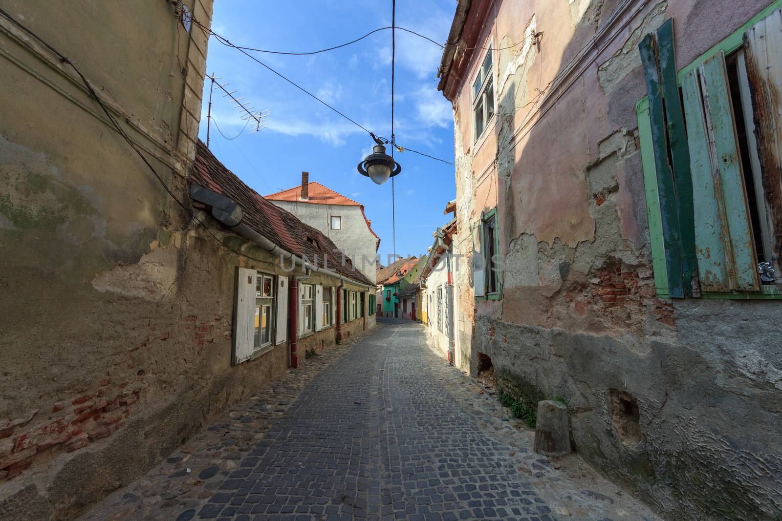 SIBIU, ROMANIA - Circa 2020: Old medieval town brick wall with cloudy blue sky. Beautiful tourist spot in eastern central Europe. Narrow old street with damaged houses. Damaged house concept. by dugulan
