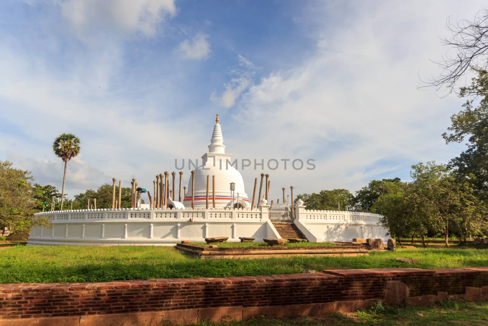 Panoramic view of restored Thuparamaya Dagoba with white bell shaped Stupa, altars, tilted vatadage pillars and Buddhist flags, Anuradhapura, Sri Lanka. Beautiful cloudy sky landscape. by dugulan