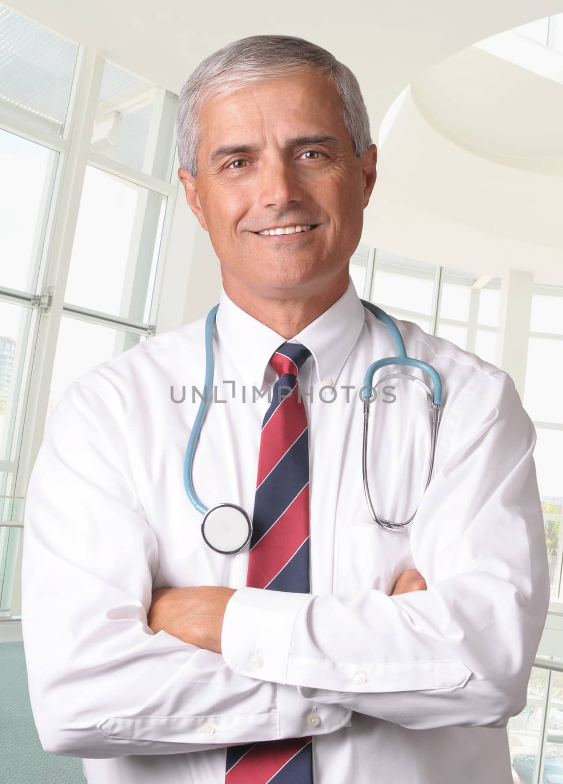 Smiling male doctor in scrubs with a stethoscope around his neck and his arms crossed. Vertical format torso view in modern looking medical facility.