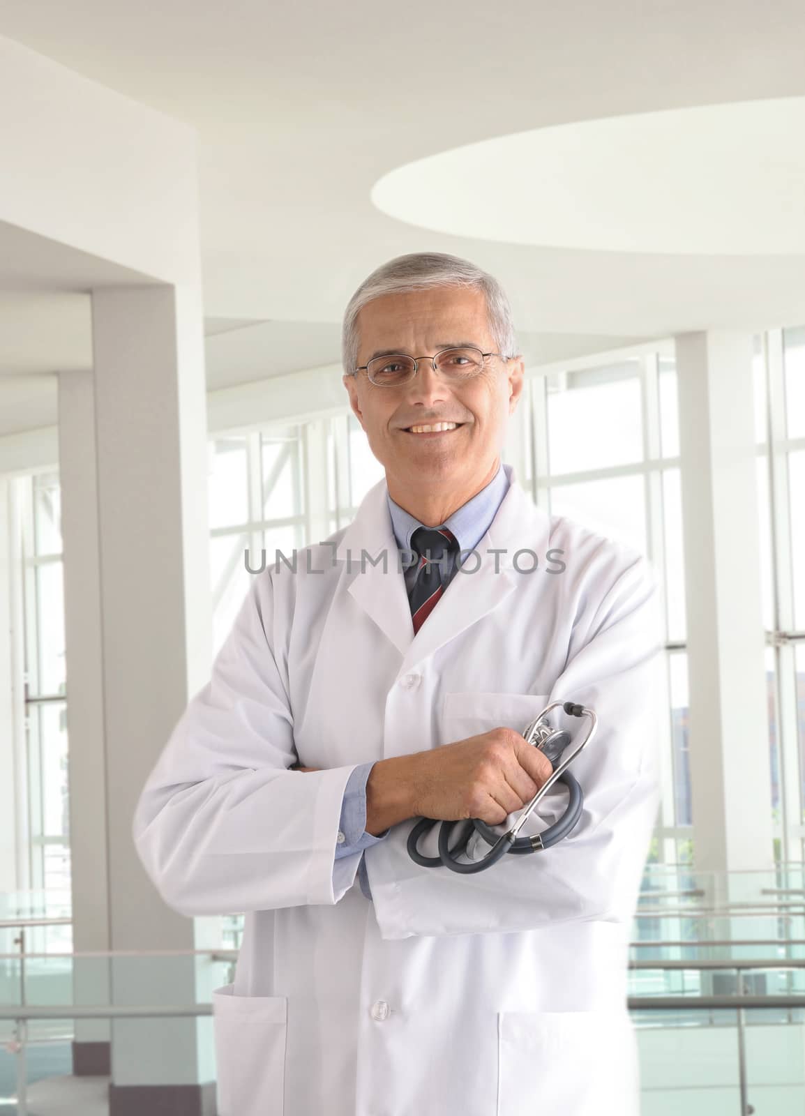 Middle aged doctor holding stethoscope with arms crossed in modern medical facility