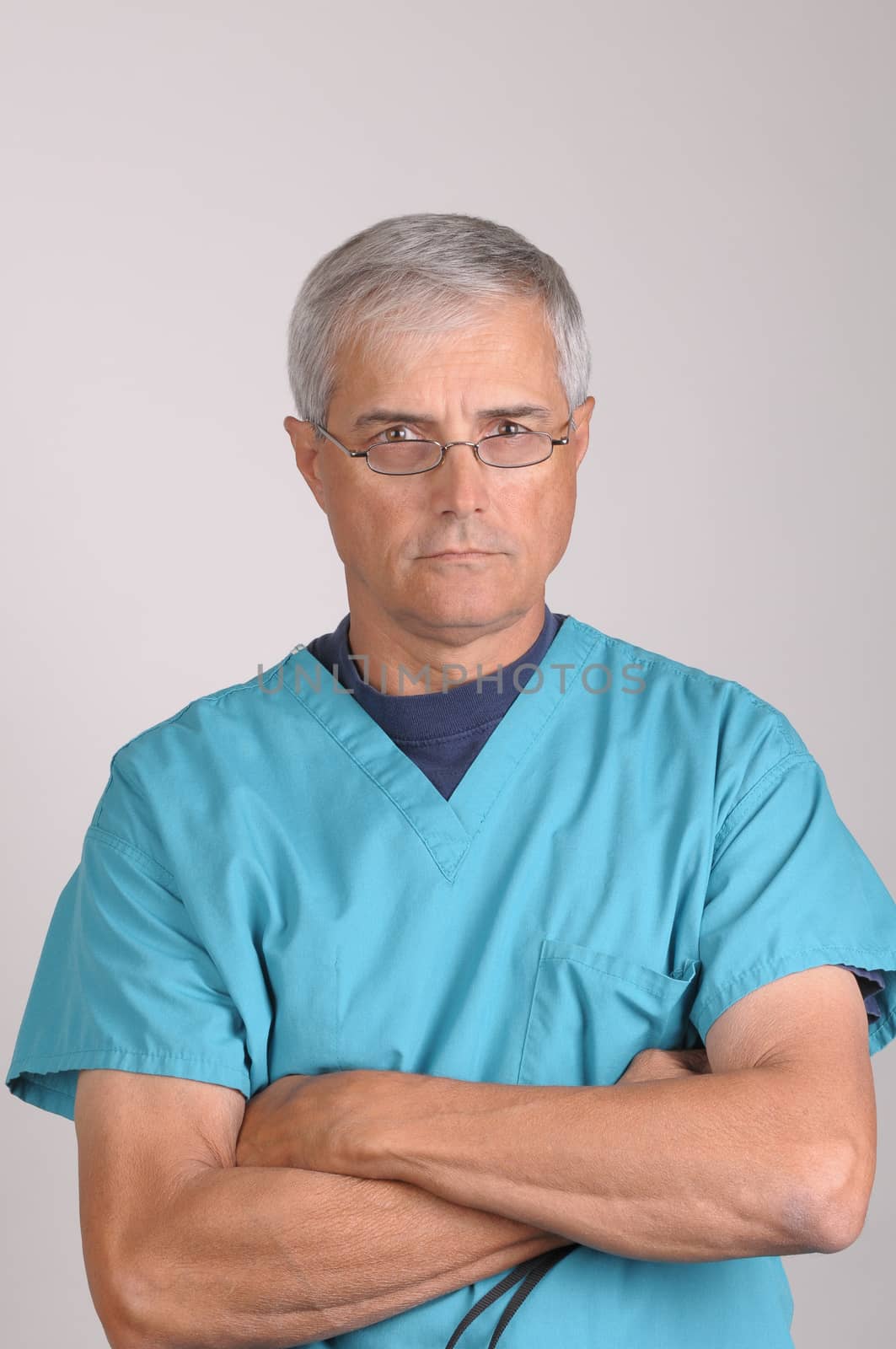 Middle Aged  Doctor in Scrubs with stern expression and his arms folded - vertical on Gray Background