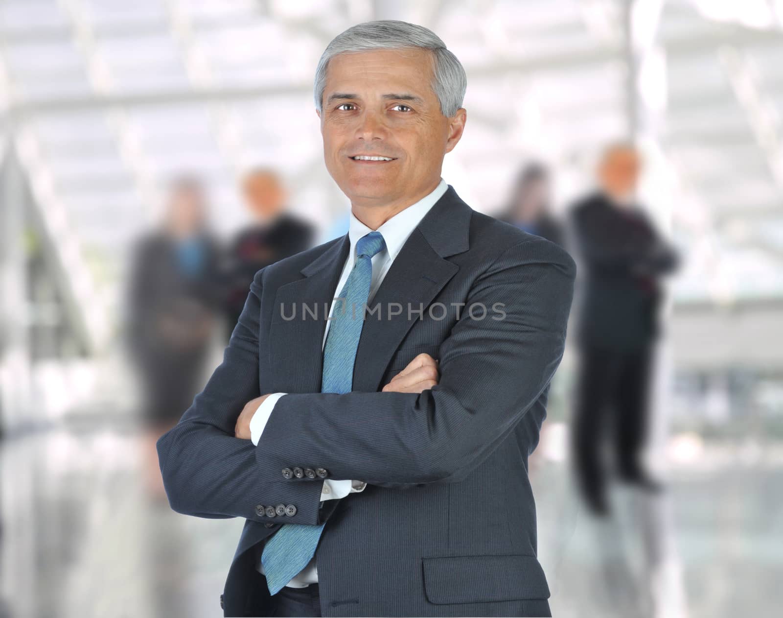 Smiling middle aged businessman in a suit and tie standing in a modern office building with his arms crossed in front of out of focus team. Square Format
