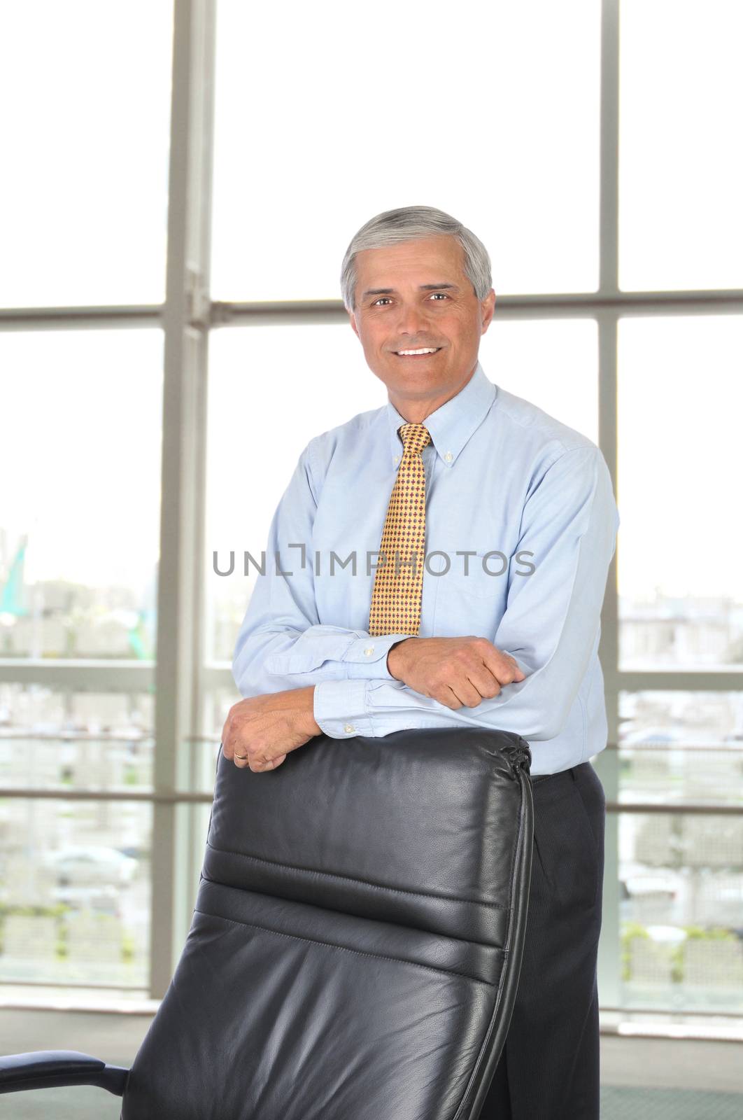 Middle aged businessman leaning on the back of his chair in office setting, Standing in front of a large window. Vertical Format