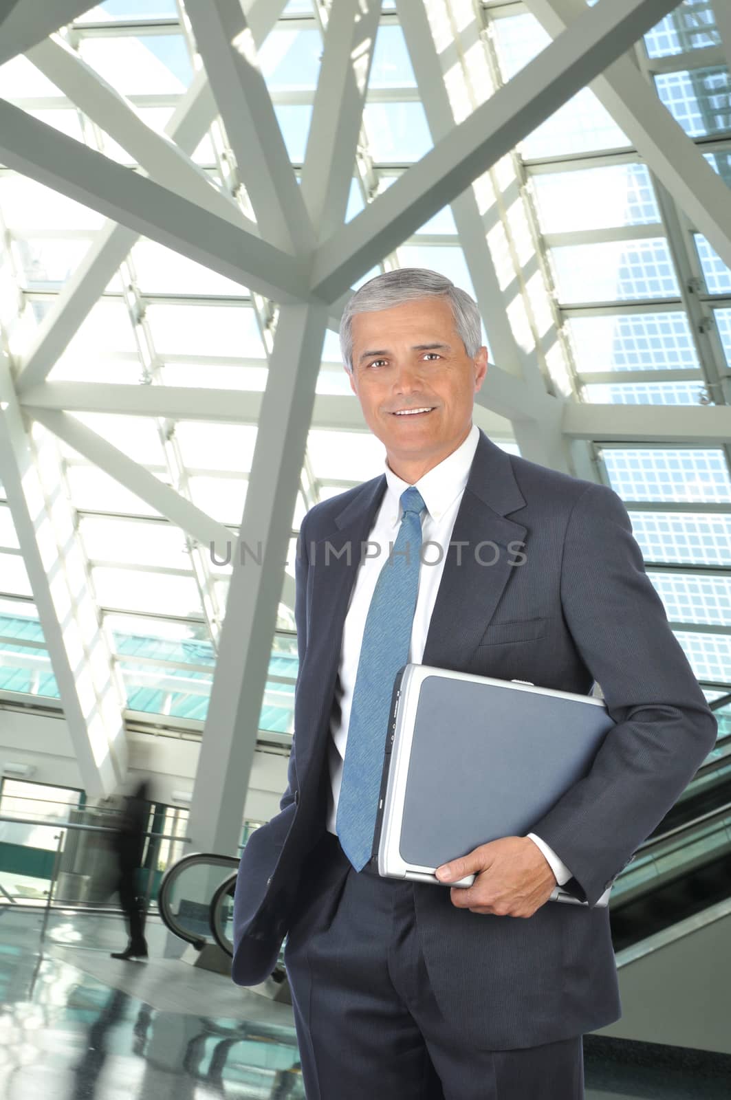 Smiling Middle aged Businessman Holding Laptop Computer Under His Arm in Building Lobby