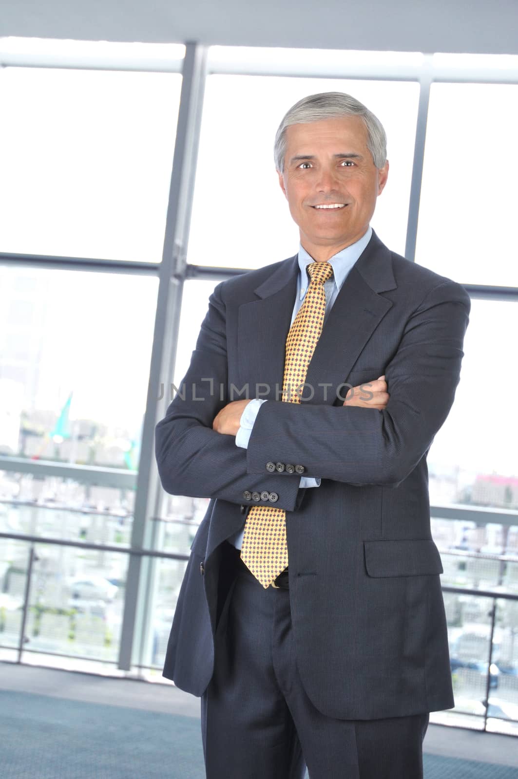 Smiling Middle aged Businessman in dark suit standing with his arms crossed in office setting