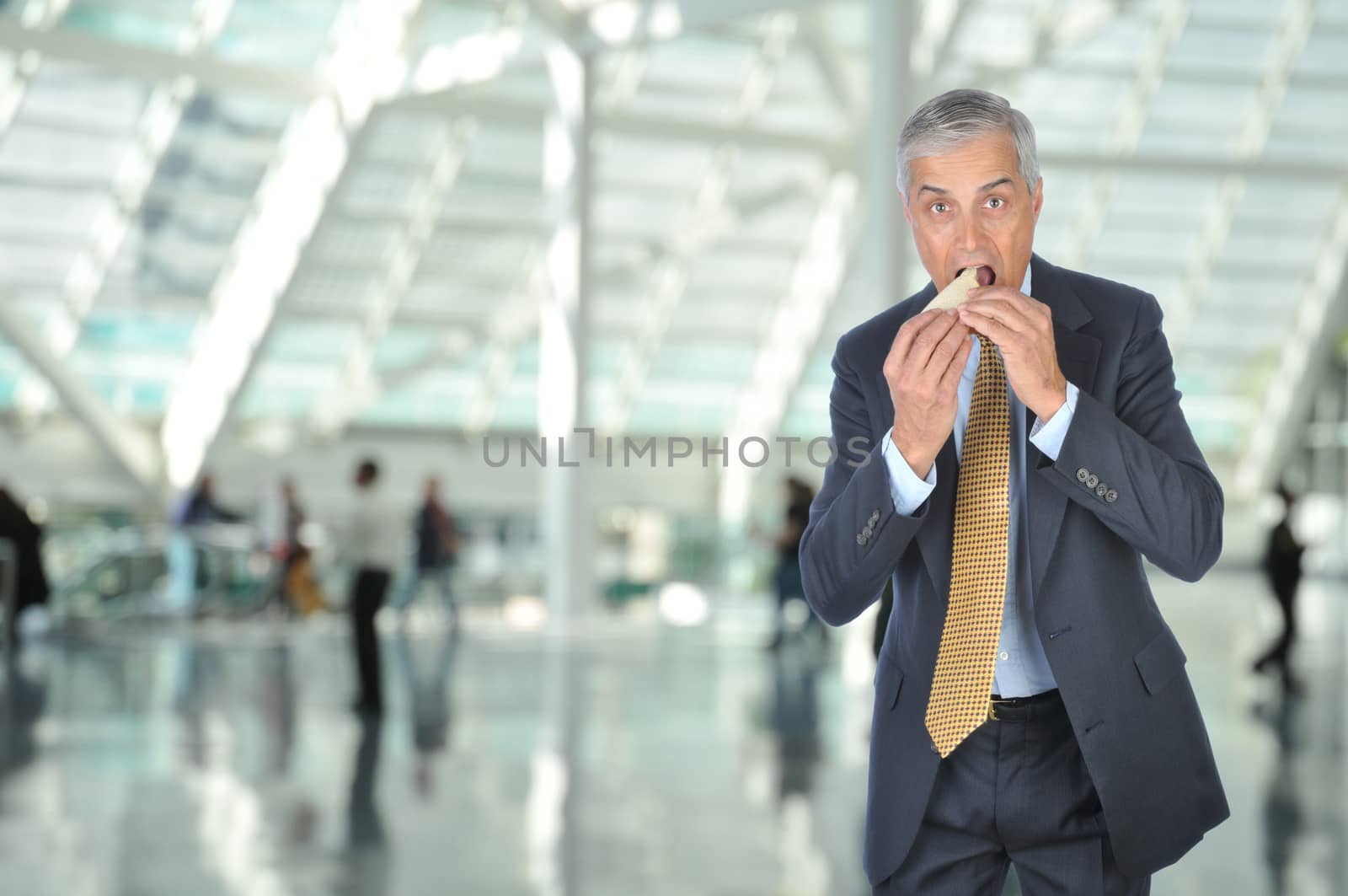 Middle aged Businessman on the go eating a sandwich in Airport Concourse, horizontal format with unrecognizable people in the background.