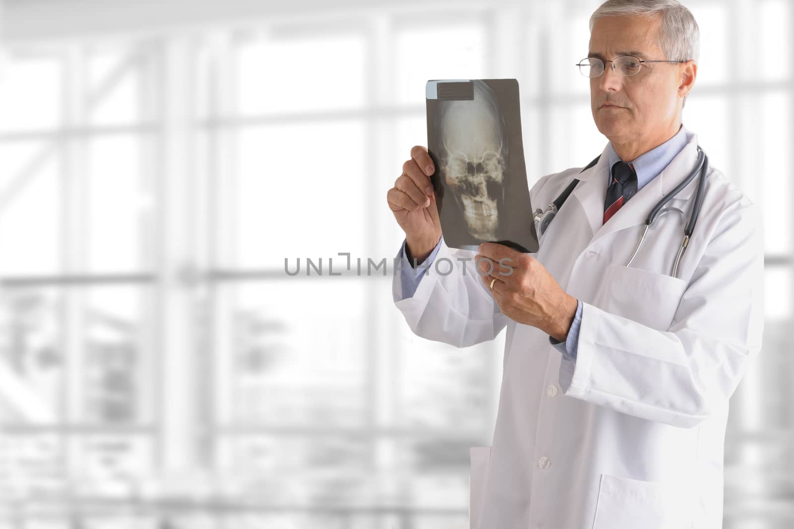 A serious senior doctor standing against office window background while looking at an X-ray.