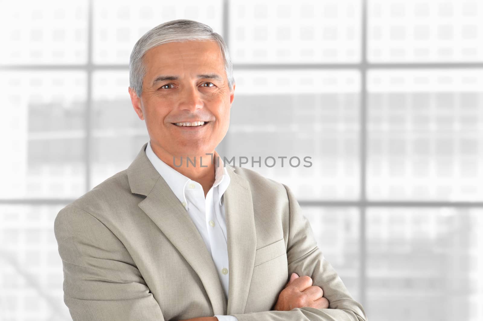 Portrait of a middle aged businessman in front of office window. Man is smiling and has his arms folded. Horizontal format.