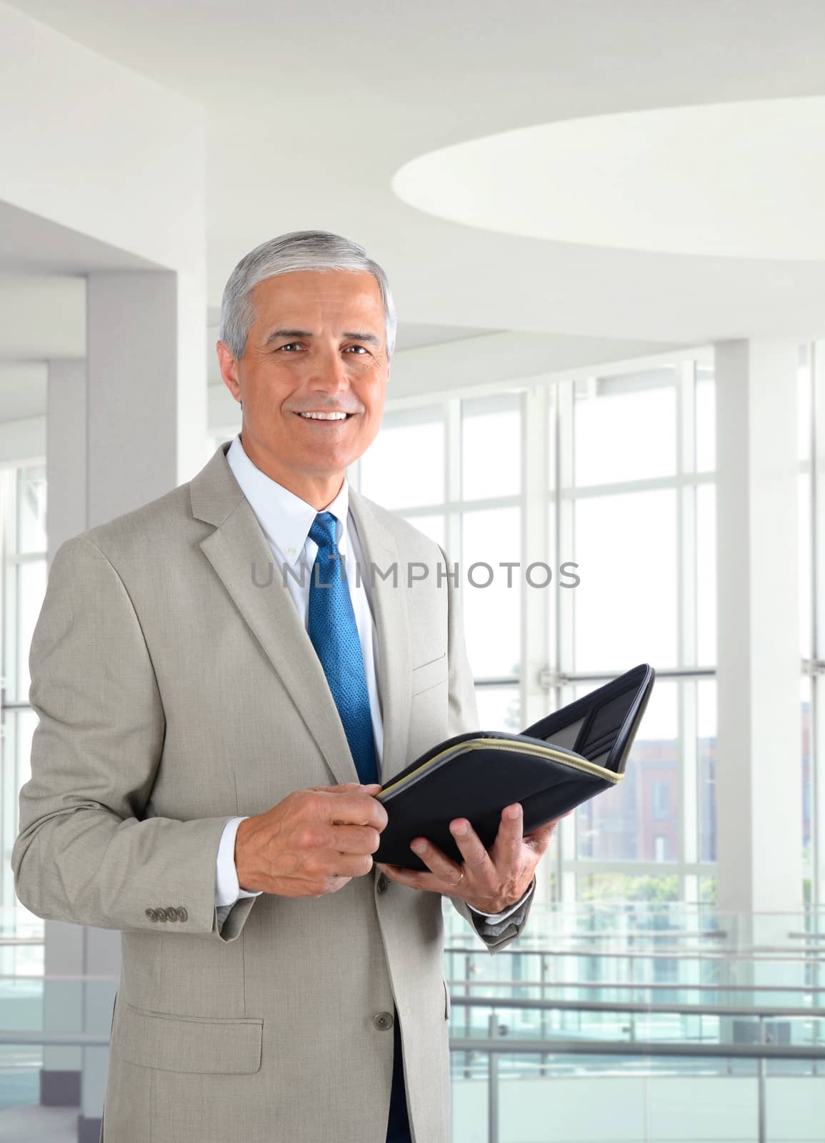 Portrait of a middle aged businessman standing in a modern office. Man is holding a small binder and smiling at the camera.