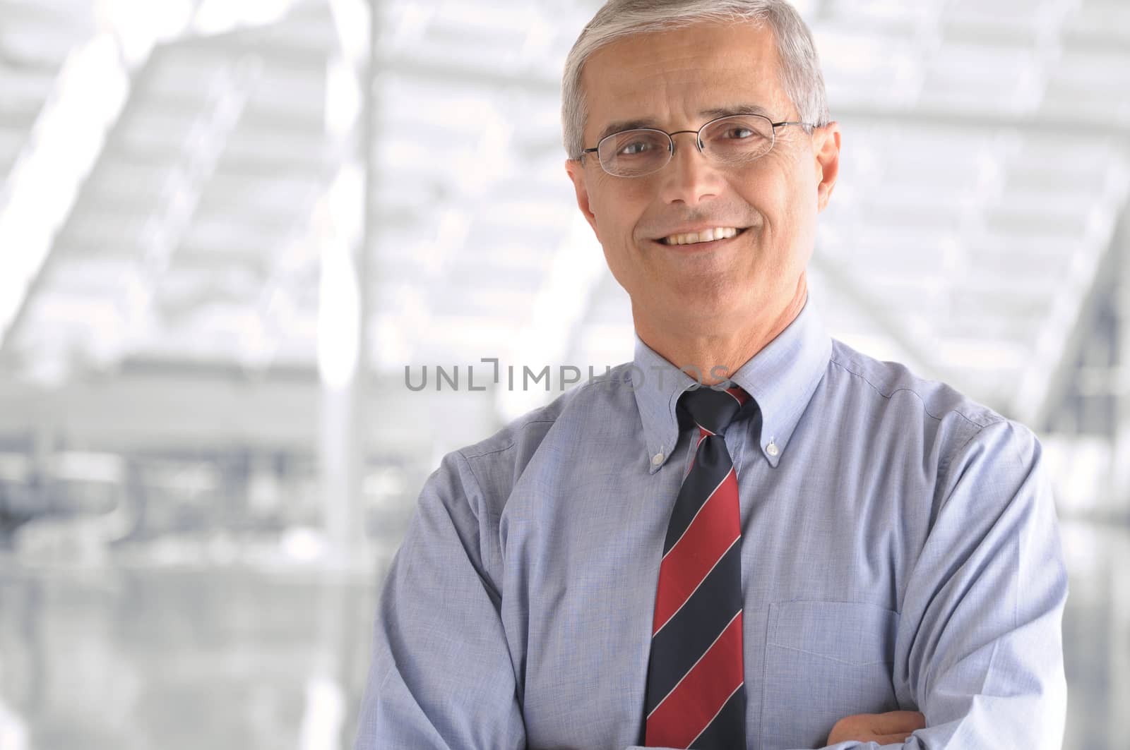 Business man portrait in modern office building. Middle aged man is smiling at the camera and has his arms folded. Closeup head and shoulders only.