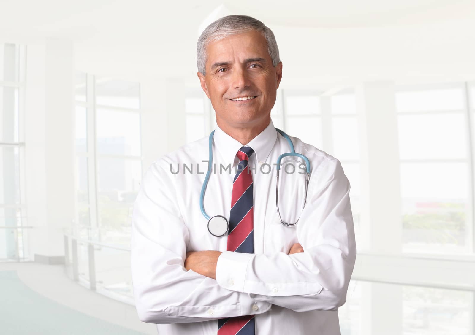 Smiling male doctor in scrubs with a stethoscope around his neck and his arms crossed. Vertical format torso view with high key background.
